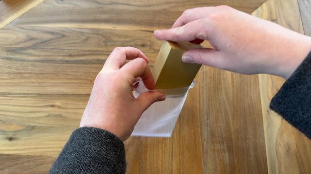 Hands place a small cardboard jewelry gift box into a sheer organza drawstring bag on a wooden table. Jewelry gifting and presentation concept.