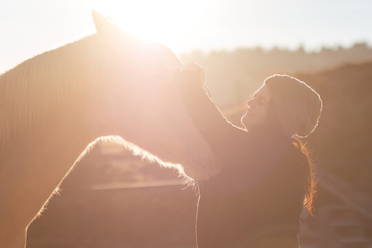 Young woman gently touching her horse outdoors, backlit by golden sunlight, calm rural atmosphere - Powered by Adobe
