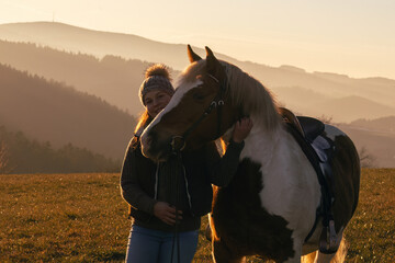 Woman gently holding her pinto horse outdoors in winter light, warm sunset mood and peaceful rural landscape