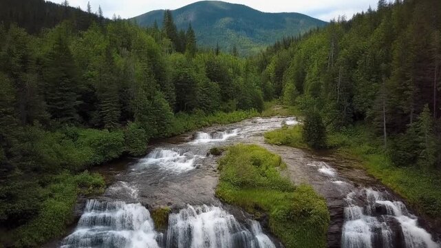 A scenic aerial view of multi tiered waterfall cascading over rocky steps through vibrant green forest with mountains and dense trees forming dramatic natural landscape