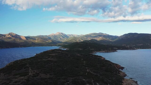 Aerial view of a rugged coastline and mountains near Cargese, Corsica, under a partly cloudy sky. The scene features dramatic landforms and deep blue sea.