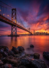 Brilliant hues of orange, pink, and purple fill the sky as the sun sets behind the city&rsquo;s skyline. Beneath the bridge, calm waters reflect the stunning spectacle, creating a magical ambiance.