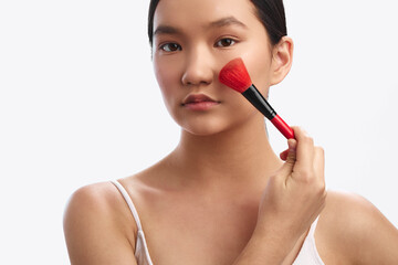 asian woman applies makeup with a cosmetic brush on her cheek in a bright studio, her calm expression and precise stroke conveying beauty, self care, and confident personal grooming.