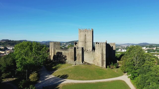 Aerial view of the historic Guimaraes Castle surrounded by greenery under a clear blue sky in Portugal. The medieval fortress stands prominently with its stone walls and towers.