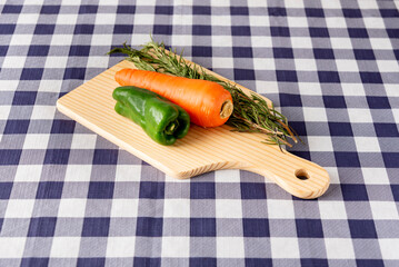Fresh bell pepper, carrot, and rosemary on a wooden cutting board over a blue checkered tablecloth. Healthy food, cooking, and preparation.