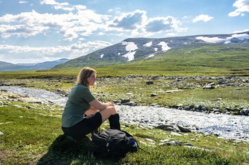A female hiker sits by a river in Trollheimen mountain area in Norway during summer. She wears zip off trousers with the pant legs are unzipped and resting on her hiking boots.

