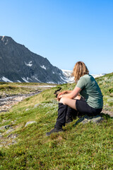 A woman sits by a river next to a hiking trail surrounded by Trollheimen mountains in Norway. She enjoys the outdoor scenery and takes a break in the summer sun