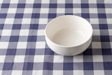 Empty white ceramic bowl on a blue and white checkered tablecloth with copy space on the left. Minimalist tableware for cereal or soup