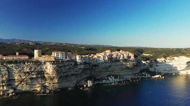 Aerial view of Bonifacio perched on dramatic white limestone cliffs above the Mediterranean Sea in Corsica, France. Picturesque historic buildings overlook the rugged coastline and clear blue water.