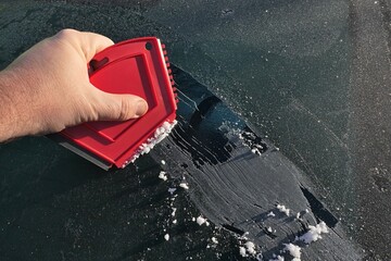 Hand of adult male person scratching down frost cover from rear window of modern C-segment sedan car with multi purpose compact red plastic scraper. 
