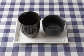 Two different black ceramic cups on white rectangular plate over blue checkered tablecloth