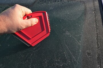 Hand of adult male person ready to remove frost cover from rear window of modern C-segment sedan car with multi purpose compact red plastic scraper. 