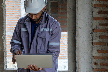 Construction foreman in safety gear stands at an unfinished building, thoughtfully reviewing plans on a laptop, illustrating decision-making, technology and supervision on a modern construction site.