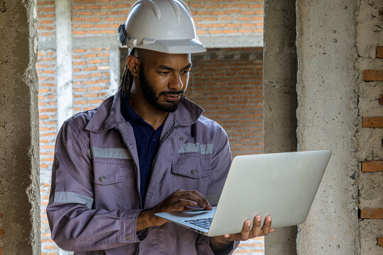Construction foreman in safety gear stands at an unfinished building, thoughtfully reviewing plans on a laptop, illustrating decision-making, technology and supervision on a modern construction site. - Powered by Adobe