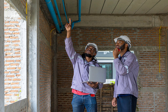 Two Black construction workers in safety helmets review plans on a laptop at a brick building site, highlighting teamwork, technology and workplace diversity in the construction industry. Team work