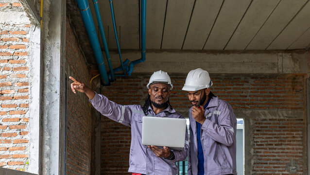 Two Black construction workers in safety helmets review plans on a laptop at a brick building site, highlighting teamwork, technology and workplace diversity in the construction industry. Team work