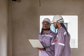 Two Black construction workers in safety helmets review plans on a laptop at a brick building site, highlighting teamwork, technology and workplace diversity in the construction industry. Team work