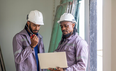 Two Black construction workers in safety helmets review plans on a laptop at a brick building site, highlighting teamwork, technology and workplace diversity in the construction industry. Team work
