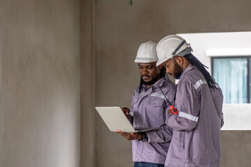 Two Black construction workers in safety helmets review plans on a laptop at a brick building site, highlighting teamwork, technology and workplace diversity in the construction industry. Team work