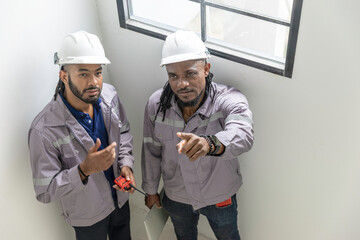 Two African construction supervisors in safety gear stand indoors, pointing and discussing work while holding a radio and laptop, showing leadership, teamwork and clear communication on site.