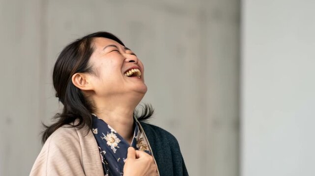 Joyful middle aged Asian woman laughing portrait smiling with hand chest wearing floral scarf layered cardigan casual blouse relaxed indoor adult