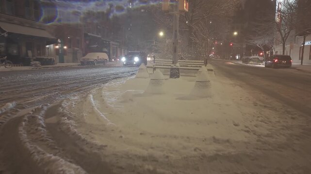 Cinematic low-altitude drone shot gliding through a snow-covered urban intersection in New York City at night. The scene features fresh snow accumulation on streets, sidewalks, and park benches, il...