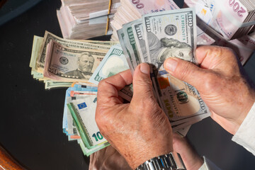A close-up image of a person counting US dollar bills. In the foreground are 100 US dollar bills,...