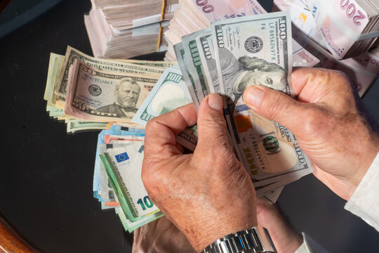 A close-up image of a person counting US dollar bills. In the foreground are 100 US dollar bills, and in the background are dollars of various denominations, Euros, and 200 Turkish Lira banknotes.