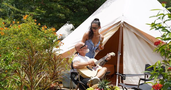 A man plays guitar for a woman near a cozy tent at a glamping site.