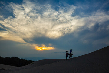Cham women walking home across the sand dunes at sunset, beneath a blue sky filled with soft white clouds.