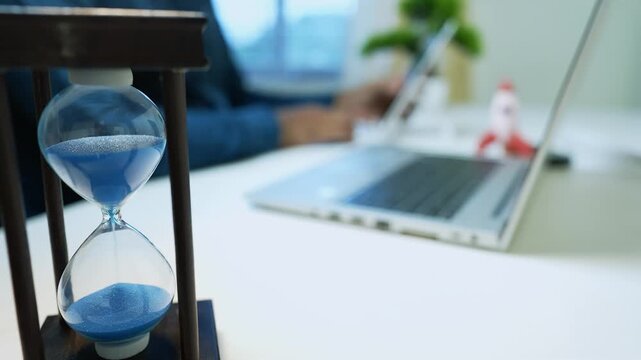 Hourglass with blue sand on desk near laptop, blurred background