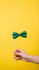 Hand Holding Green Glitter Bow Tie Prop on Stick for St. Patrick's Day Celebration over Yellow Background