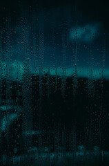 Raindrops on a glass windowpane, with a dark and moody urban background into bokeh. Deep blue and...