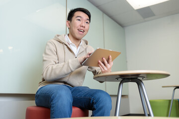 A Chinese student smiles while holding a tablet at his school desk. He appears engaged with digital learning technology in a bright modern classroom setting.