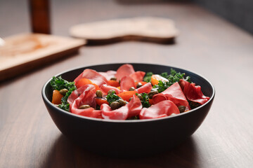 Salad with kale, bresaola and cherry tomatoes decorated with capers in black bowl on walnut wood table