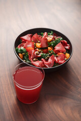 Salad with kale, bresaola and cherry tomatoes decorated with capers in black bowl and glass of red drink on walnut wood table