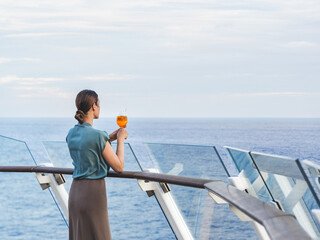 Stylish woman with a glass of drink standing on the empty deck of a cruise liner against the backdrop of sea waves. Sunny day. Chill lifestyle, luxury travel. Perfect for holiday and travel themes