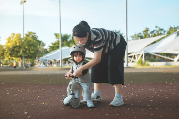 Asian mother helping and teaching her little toddler daughter to ride a kick scooter with a safety helmet at an outdoor park for family activity concept
