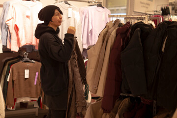 Woman inspecting winter coats on rack, focused bargain hunter checks fabric and price tags while evaluating fit, warm store lighting, layered displays, crowded aisle, casual outerwear