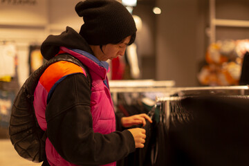 shopper comparing texture on clothing rack, close inspection of knit and weave, tag and price visible, beanie and puffer vest worn, tactile evaluation for quality and style choice in store