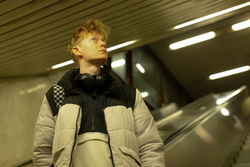 young caucasian man near escalator subway wearing puffer jacket and headphones, gazing upward with thoughtful expression, fluorescent lighting casting warm highlights, modern underground
