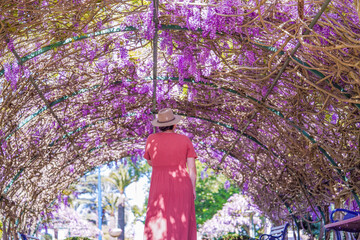 Woman in Hat and Coral Dress Standing Under Archway of Blooming Purple Wisteria Flowers
