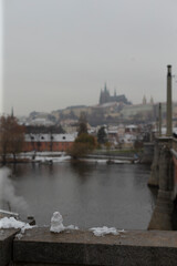 Obraz premium Prague bridge parapet with fresh snow, stone balustrade dusted with snowball remnants, distant castle silhouette across Vltava, single commuter crossing under overcast winter light, chilled