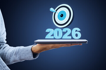 2026 target goal concept with dartboard hovering above tablet in hand representing business success and digital strategy on blue background.