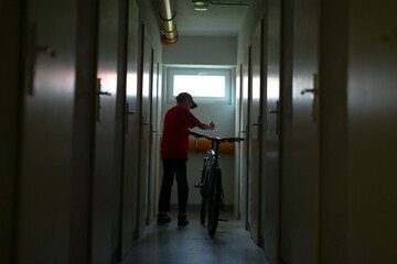 cyclist pushing bicycle down narrow corridor, red shirt and cap visible, courier vibe and gig economy energy, lowlight urban transit, helmet and gear implied, movement toward exit