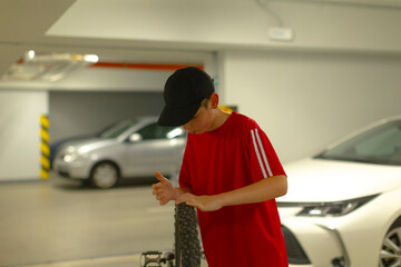 young man in underground parking checking smartphone while leaning on bicycle near parked cars, fluorescent lights casting cool shadows, casual red shirt and black cap, quiet evening