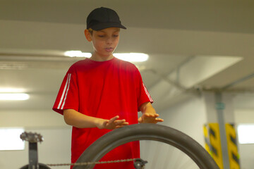 White boy checking bicycle wheel in indoor garage, red shirt and cap, focused hands on tire and spokes, concrete pillars and fluorescent lighting, casual maintenance moment before ride