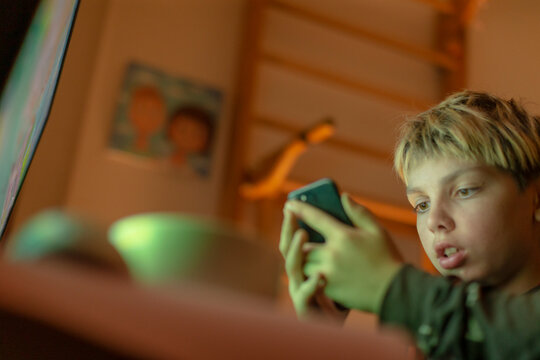 Caucasian teenage boy focused on smartphone screen dim bedroom green ambient glow gaming or scrolling with intense expression earbuds in, late night digital routine and online - Powered by Adobe