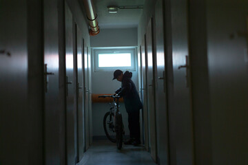young cyclist in corridor preparing bicycle, student adjusts saddle and inspects tires next to locker area under cool daylight, casual urban scene showing micromobility and daily routine