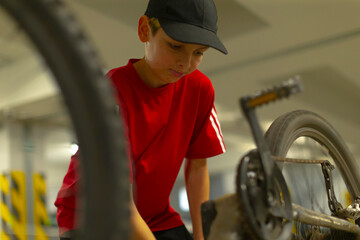 Contemplative caucasian teenage boy near bicycle, soft gaze downward, red shirt and black cap, shallow depth of field highlights emotion and texture of chain and wheel, quiet